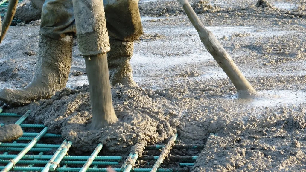Worker's rubber boots on green-coated rebar mesh in freshly poured wet concrete, with a hose and poker in the mix